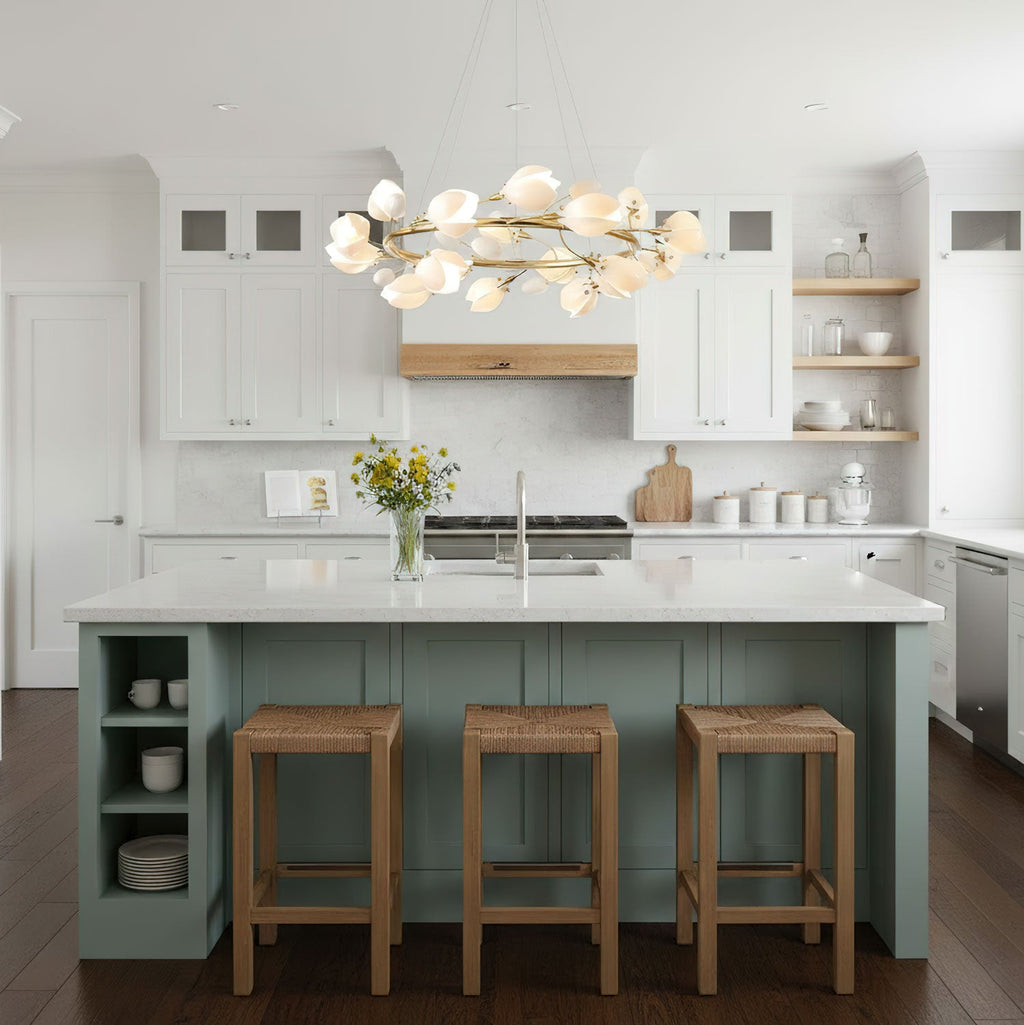 Gold-ringed Clérachic chandelier with white petals above a sage-green kitchen island with three rattan stools and open shelving.