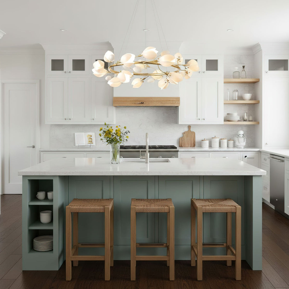 Gold-ringed Clérachic chandelier with white petals above a sage-green kitchen island with three rattan stools and open shelving.