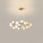 Lit Clérachic chandelier with a crowded ring of white ceramic petals, hung from a gold canopy on fine cables against a beige wall.