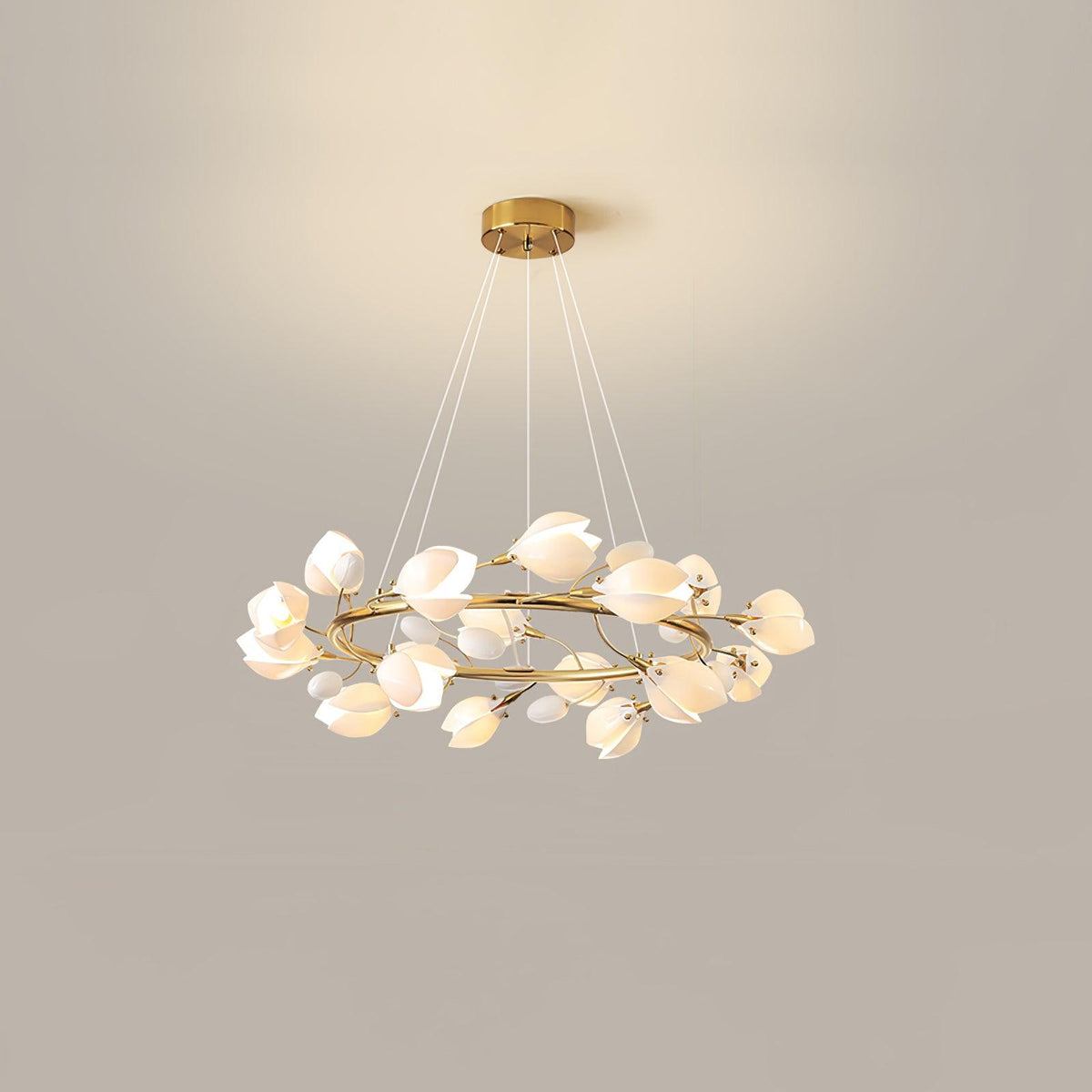 Lit Clérachic chandelier with a crowded ring of white ceramic petals, hung from a gold canopy on fine cables against a beige wall.