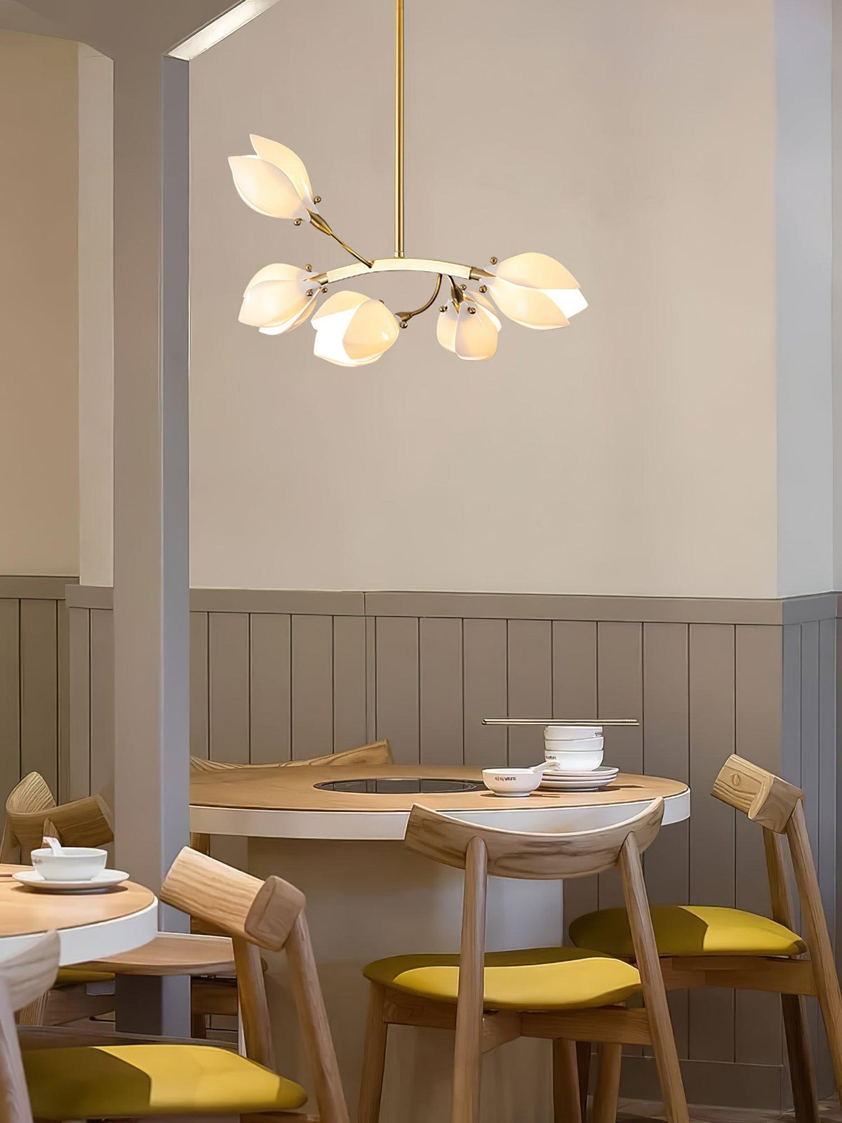 Clérachic petal chandelier on a gold rod above a round wooden table and mustard-seated chairs, beside a panelled wall.