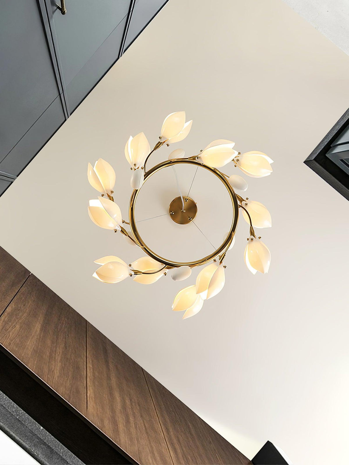Looking up at a Clérachic ring chandelier on a white ceiling, with petals circling the gold ring and a dark wood stairwell at the side.