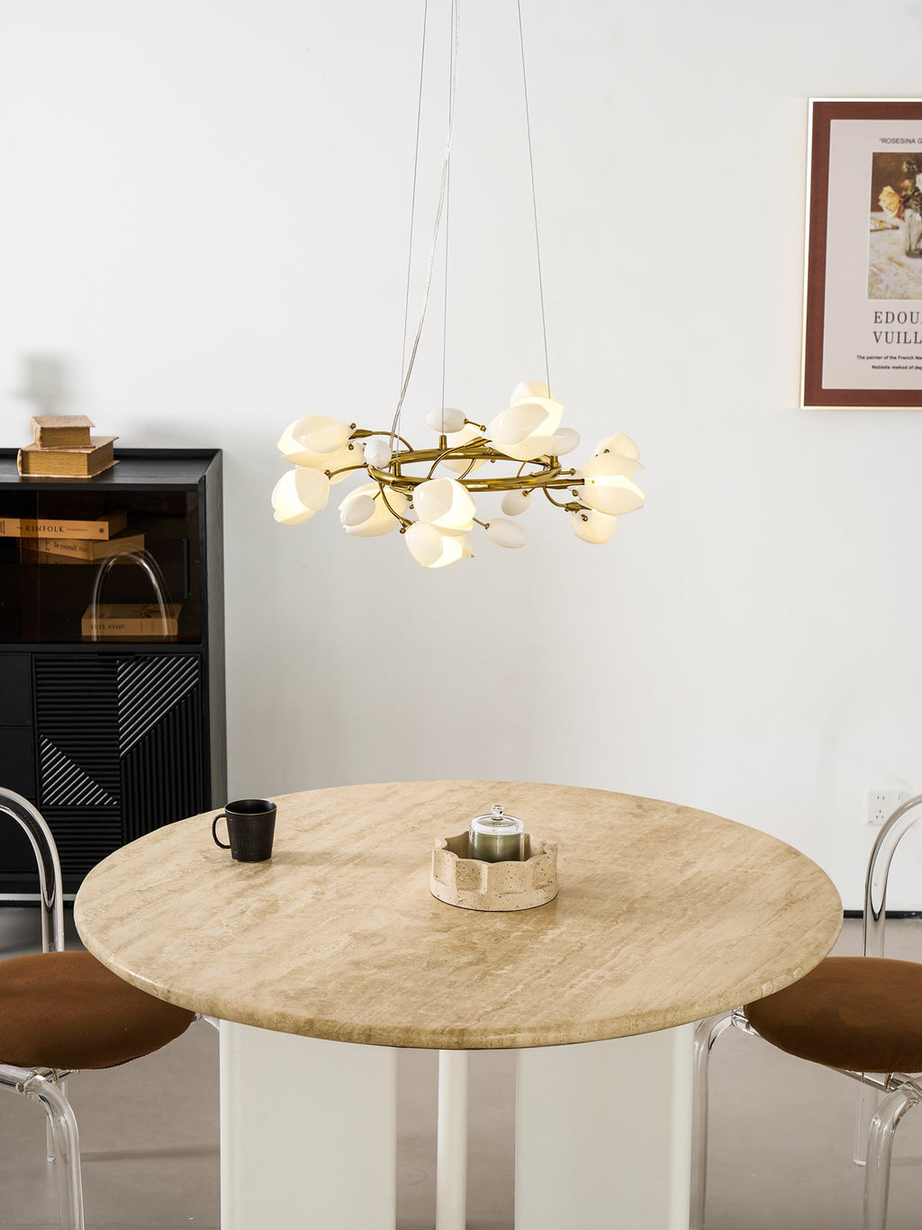 Clérachic ring chandelier lit and hung low over a round travertine table with a dark mug, near a black cabinet and a framed poster.