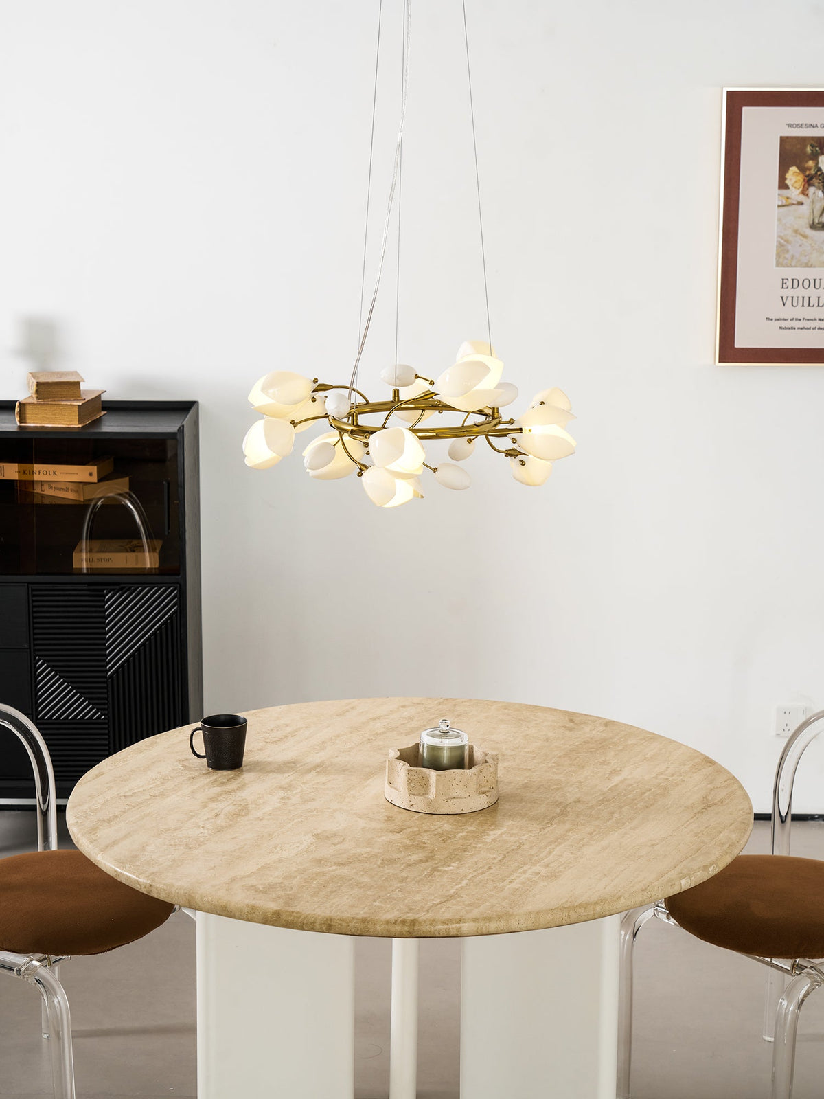 Clérachic ring chandelier lit and hung low over a round travertine table with a dark mug, near a black cabinet and a framed poster.