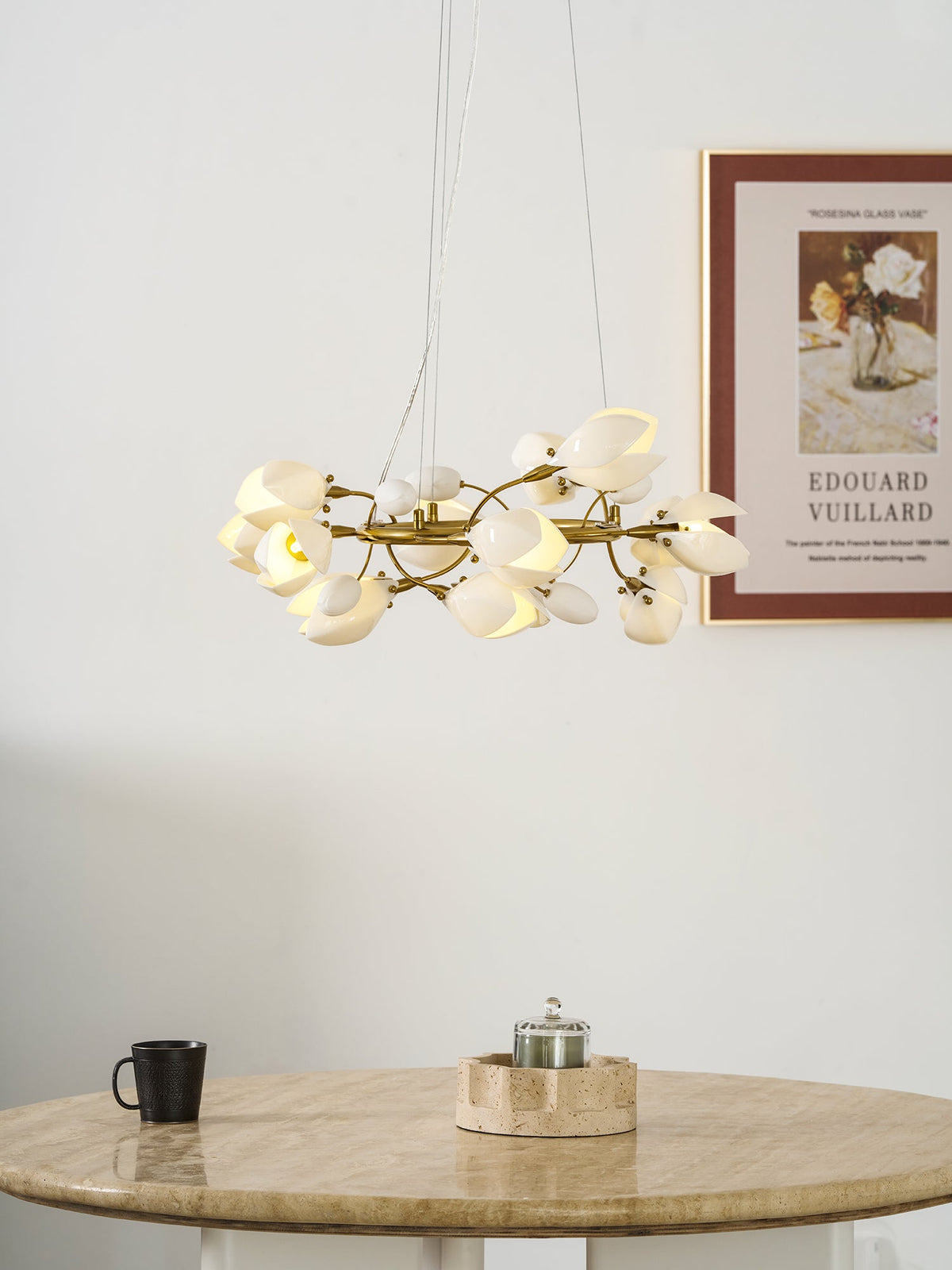 A Clérachic ring chandelier lit above a round travertine table, with a framed print on the wall and a black mug on the table.