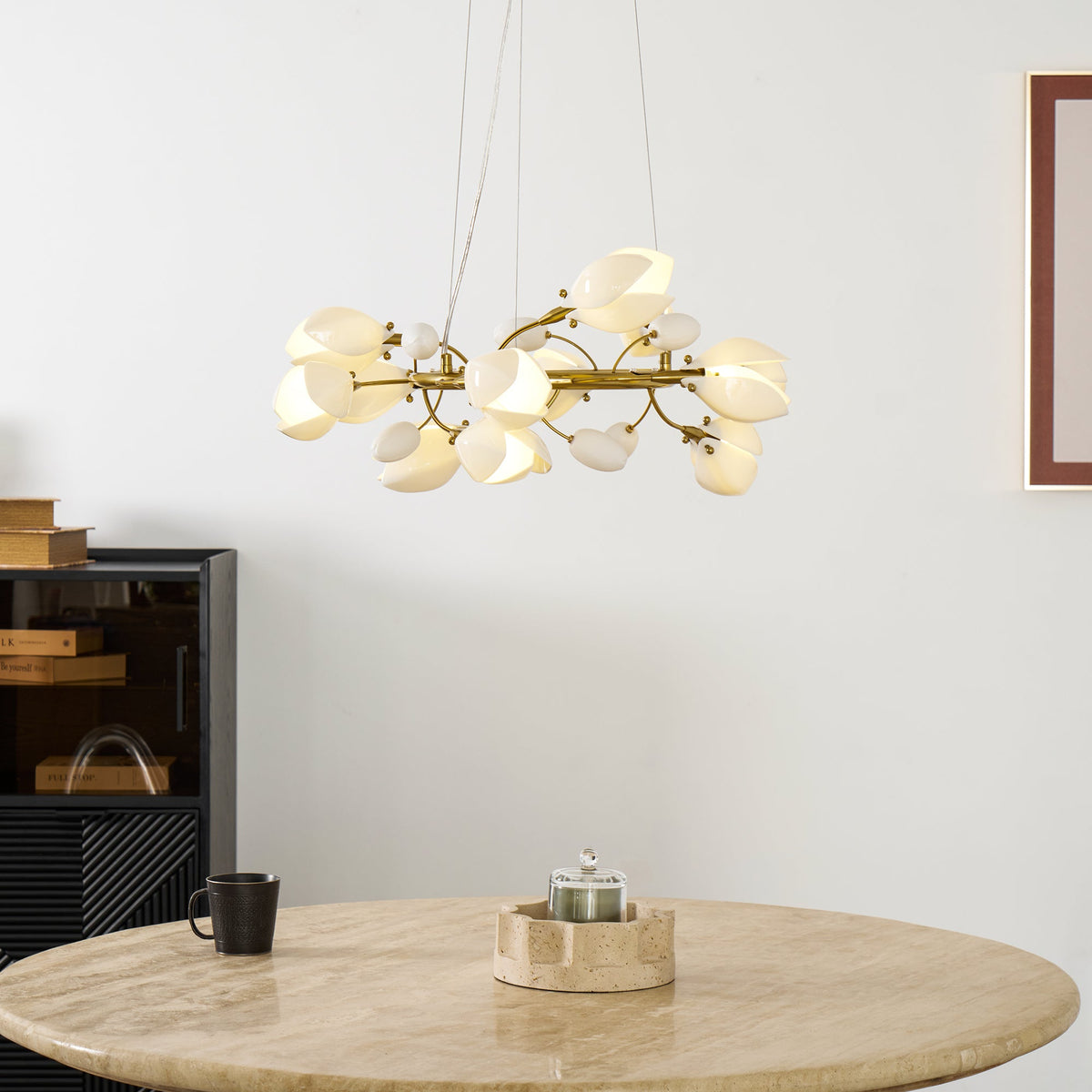 Lit Clérachic chandelier with white petals hanging above a round travertine table, a black mug and a black cabinet.