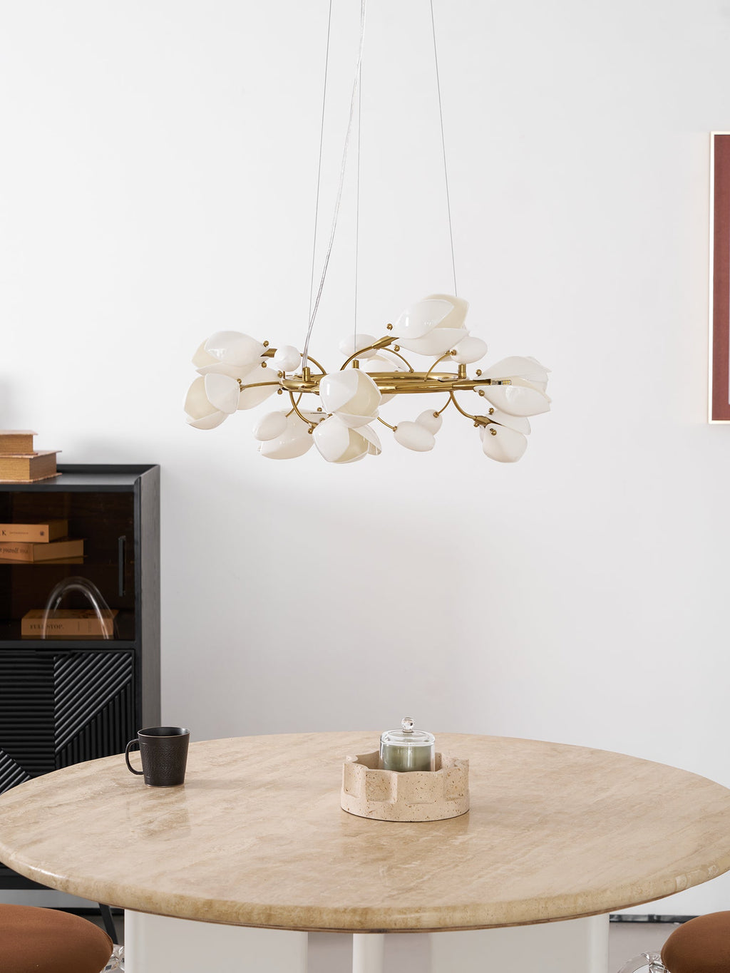 Lit Clérachic ring chandelier above a round travertine table with a candle holder and a black mug, beside a dark cabinet.