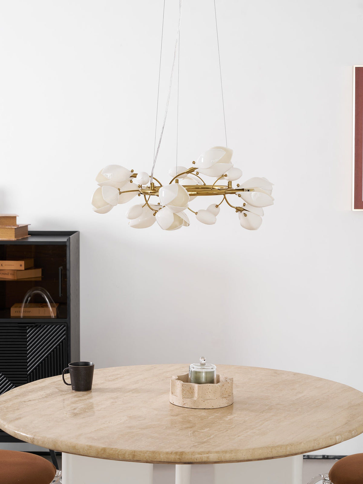 Lit Clérachic ring chandelier above a round travertine table with a candle holder and a black mug, beside a dark cabinet.