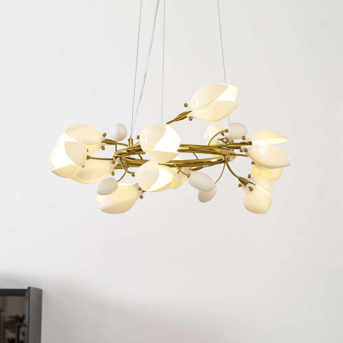 Lit Clérachic ring chandelier seen from below, with white petals and buds on twisting gold branches, against a pale wall.