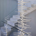 Clérachic's white bar chandelier lights a staircase, its slim bars crossing beside a glass balustrade.