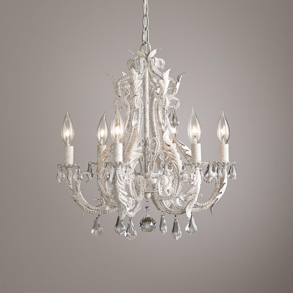 A Clérachic chandelier with white scrolling arms and clear crystal pendants, photographed against a soft mauve-grey wall.