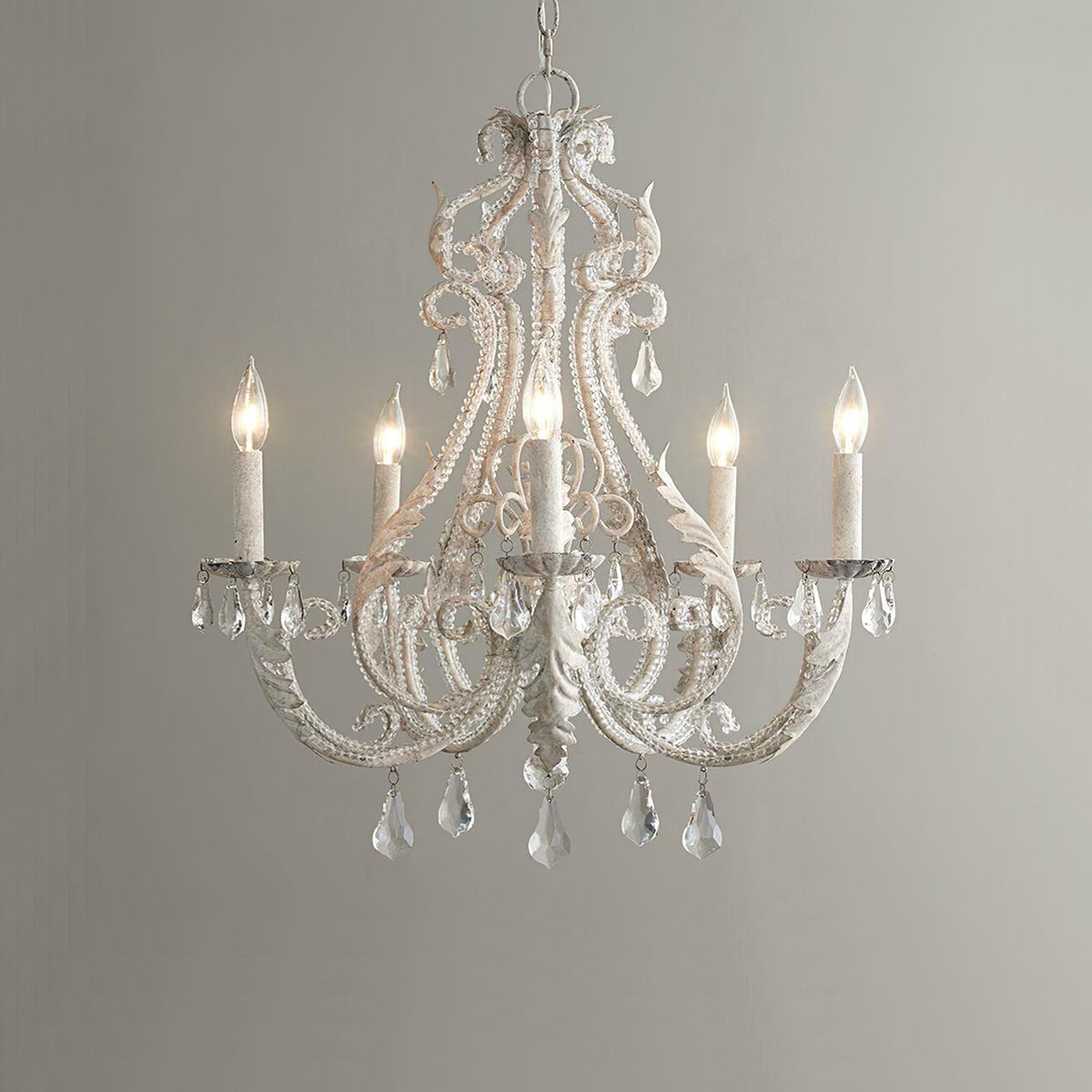 Straight-on view of a Clérachic chandelier with unlit candle sleeves, beaded scrollwork and crystal drops on a grey backdrop.