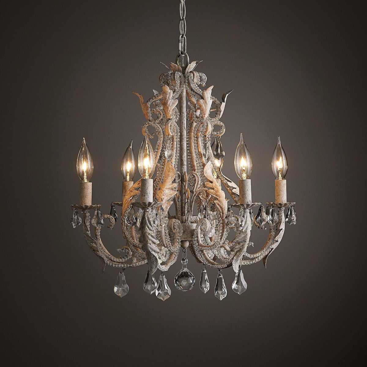 A Clérachic chandelier lit against a dark brown backdrop, with warm candle bulbs glowing behind its crystal drops.