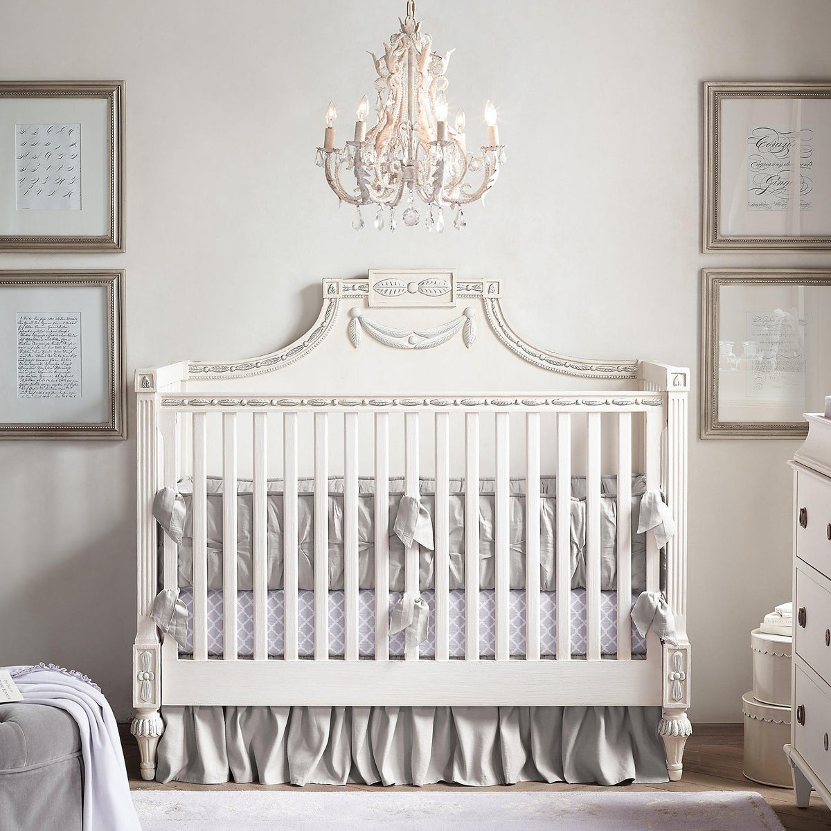 Above a white cot with grey bedding in a pale nursery, this Clérachic chandelier hangs with framed prints on the walls.