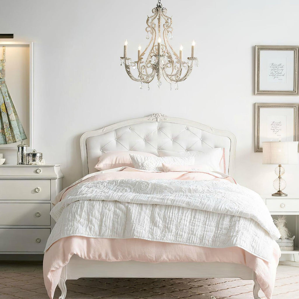 Over a white tufted bed with blush bedding, this Clérachic chandelier hangs in a pale bedroom with framed prints.