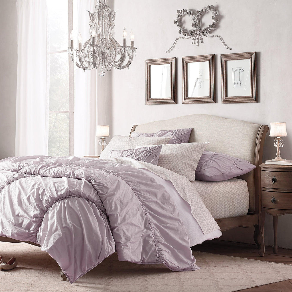 A Clérachic chandelier hangs in a bedroom with a lilac duvet and a wooden-framed headboard, with framed sketches behind.