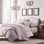 A Clérachic chandelier hangs in a bedroom with a lilac duvet and a wooden-framed headboard, with framed sketches behind.
