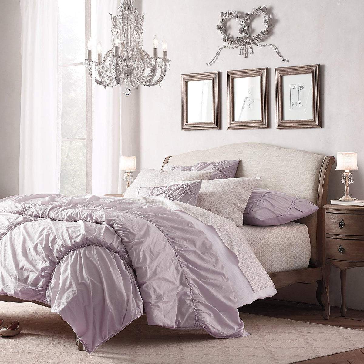A Clérachic chandelier hangs in a bedroom with a lilac duvet and a wooden-framed headboard, with framed sketches behind.