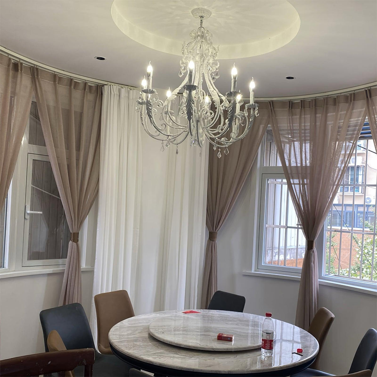 In a round dining room with sheer curtains and a marble table, this Clérachic chandelier is lit beneath a circular ceiling recess.