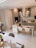 A lit white Clérachic chandelier over a kitchen island, in an open-plan room with a marble floor and a pale ceiling rose.
