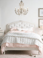 A Clérachic chandelier centred above a white upholstered bed with pink and white bedding in a bright bedroom.