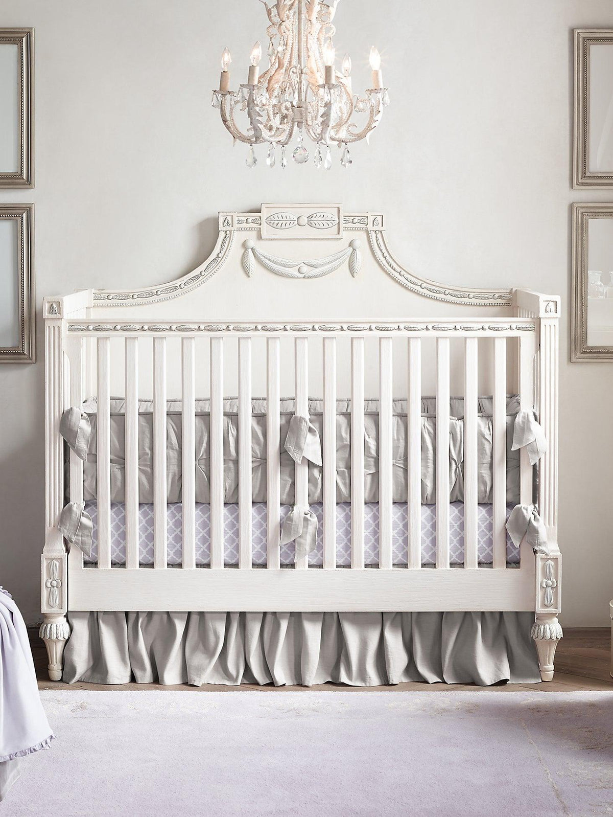 Looking up at a Clérachic chandelier hung over a white cot with grey bedding, in a pale nursery with framed prints.