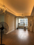 A lit Clérachic chandelier in a long, warm-lit room with a dark wooden floor, a cove-lit ceiling and a fan at the left.