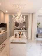 A white Clérachic chandelier lit above a white marble cube table, in a bright room with a kitchen area behind it.