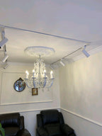 A lit Clérachic chandelier below a plaster ceiling rose in a small room with black armchairs and white wall panelling.