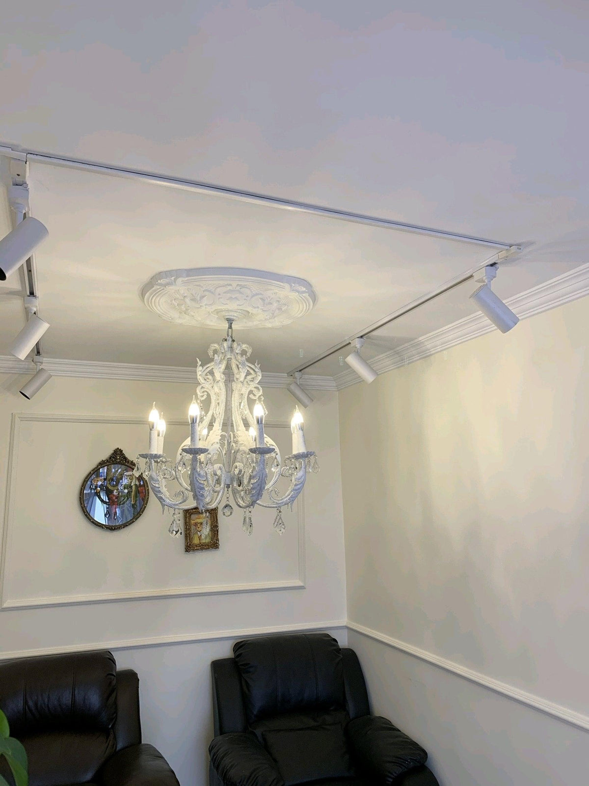 A lit Clérachic chandelier below a plaster ceiling rose in a small room with black armchairs and white wall panelling.