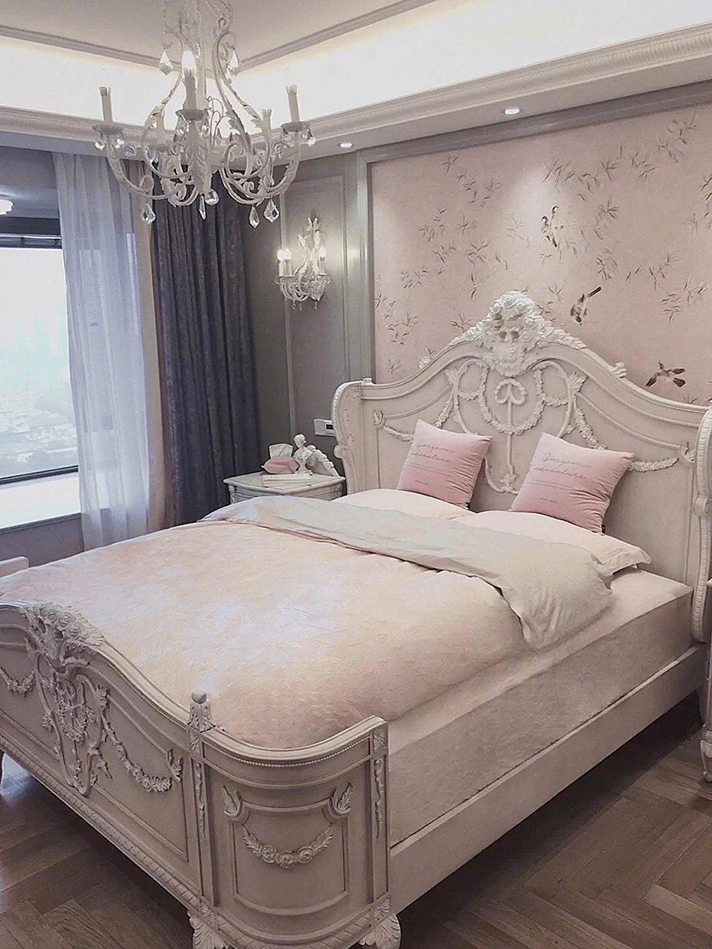 A Clérachic chandelier hangs in a pink-and-cream bedroom with a carved bed, a dark ceiling coffer and a floral wall panel.