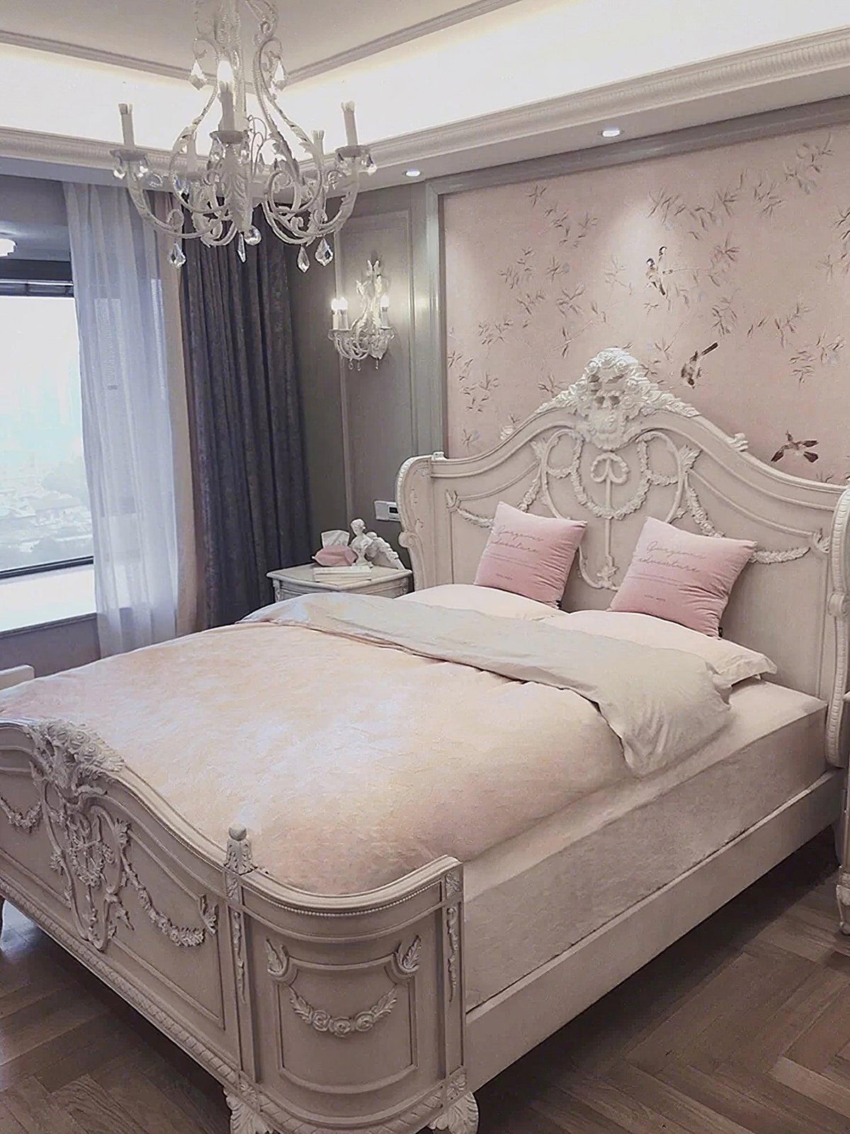 A Clérachic chandelier hangs in a pink-and-cream bedroom with a carved bed, a dark ceiling coffer and a floral wall panel.