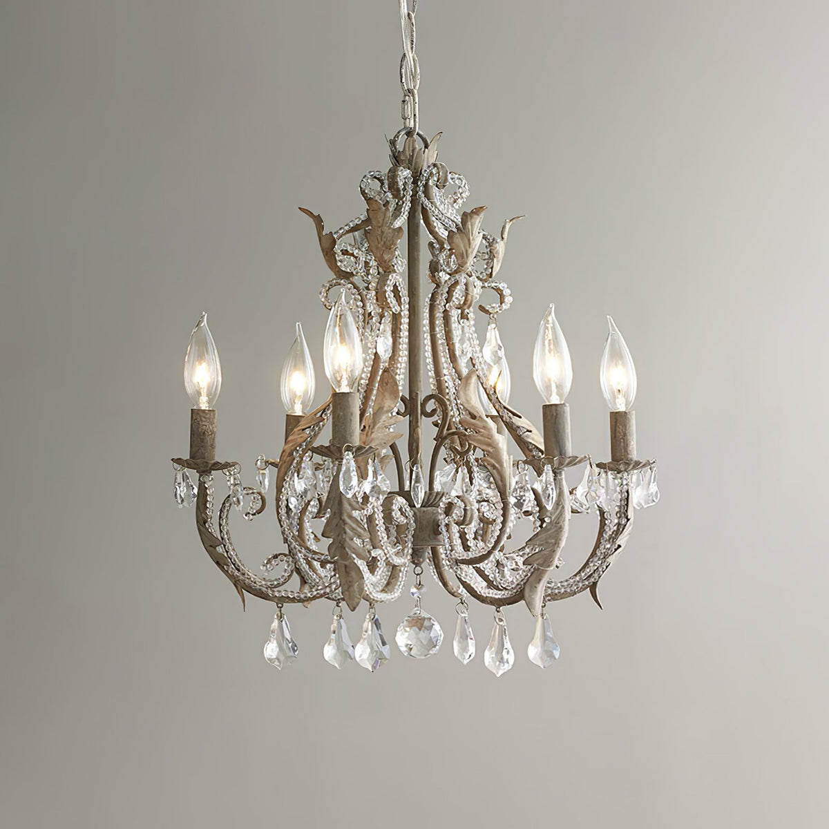 A Clérachic chandelier in an aged taupe-brown version with clear crystal drops, lit with candle bulbs against a grey wall.
