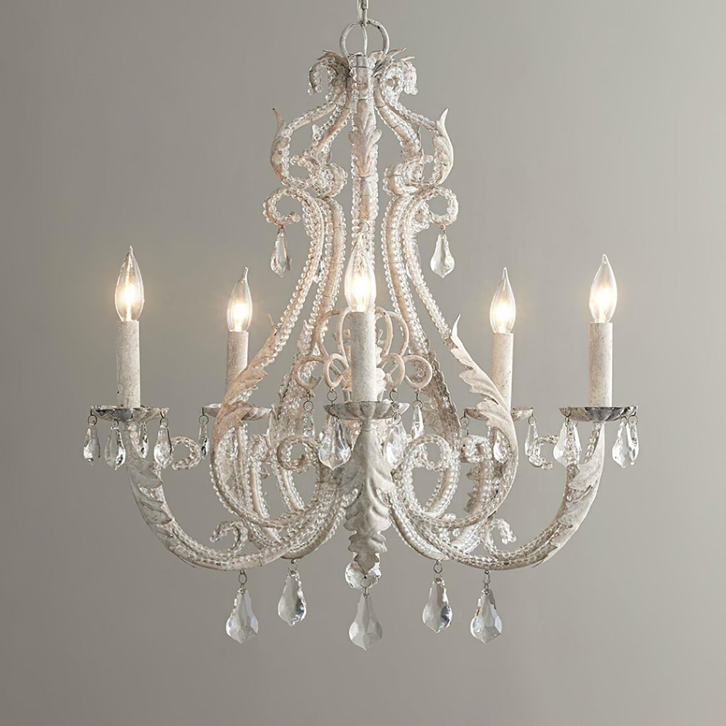 A Clérachic chandelier lit warm white against a pale grey wall, with beaded scrollwork and crystal drops hanging below.