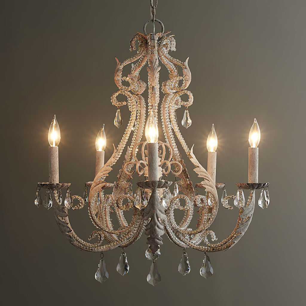 A Clérachic chandelier lit against a dark olive wall, with warm candle bulbs glowing through its weathered cream scrollwork.
