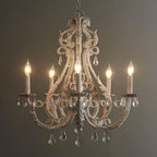 A Clérachic chandelier lit against a dark olive wall, with warm candle bulbs glowing through its weathered cream scrollwork.