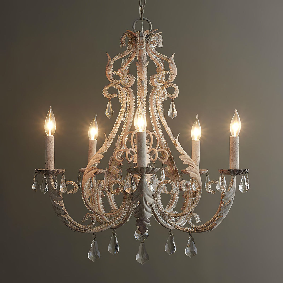 A Clérachic chandelier lit against a dark olive wall, with warm candle bulbs glowing through its weathered cream scrollwork.