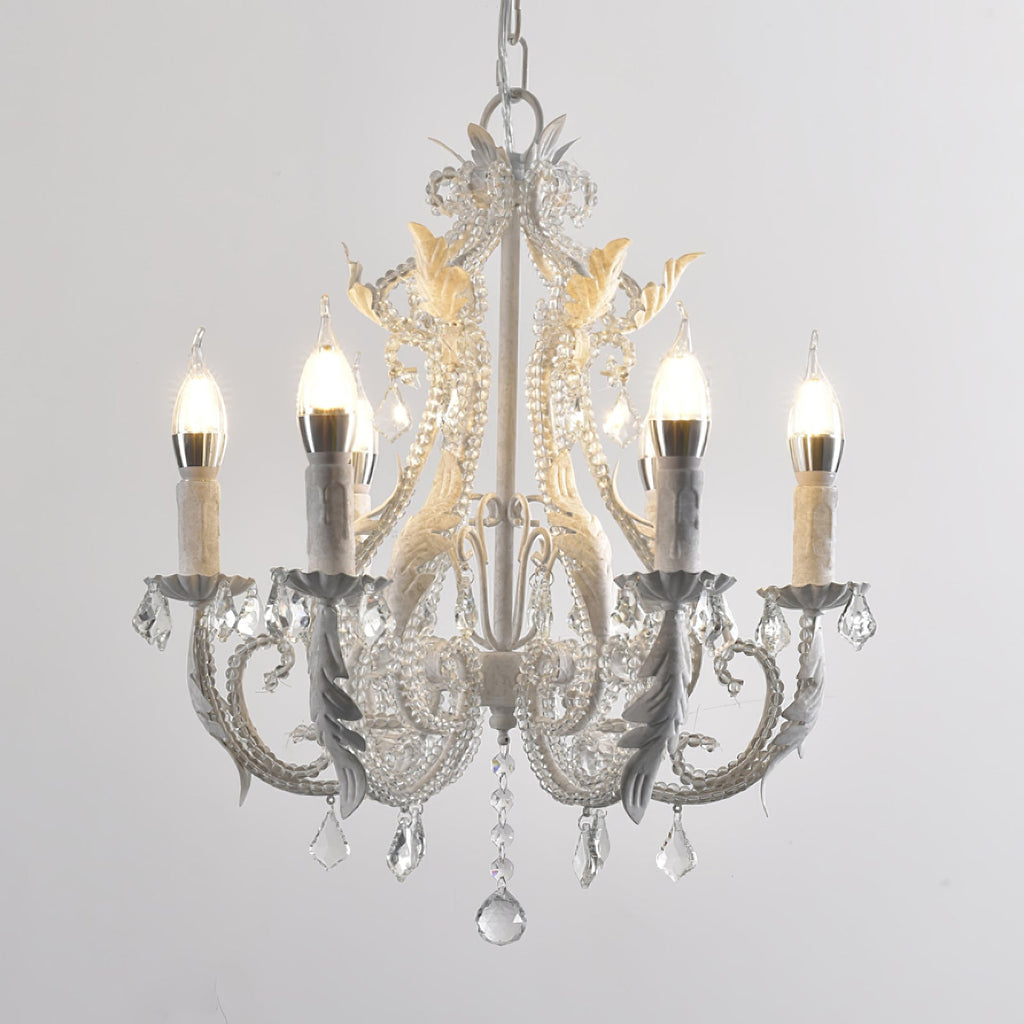 A Clérachic chandelier with larger frosted candle bulbs, white scrolled arms and a long crystal drop, on a grey backdrop.