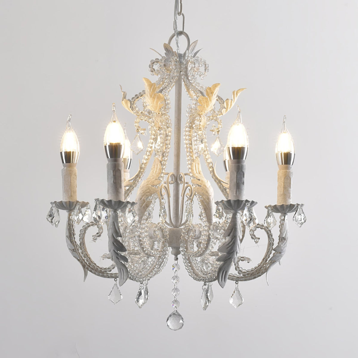 A Clérachic chandelier with larger frosted candle bulbs, white scrolled arms and a long crystal drop, on a grey backdrop.