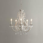 A Clérachic chandelier with unlit candle sleeves, beaded white scrollwork and crystal drops, against a mid-grey wall.