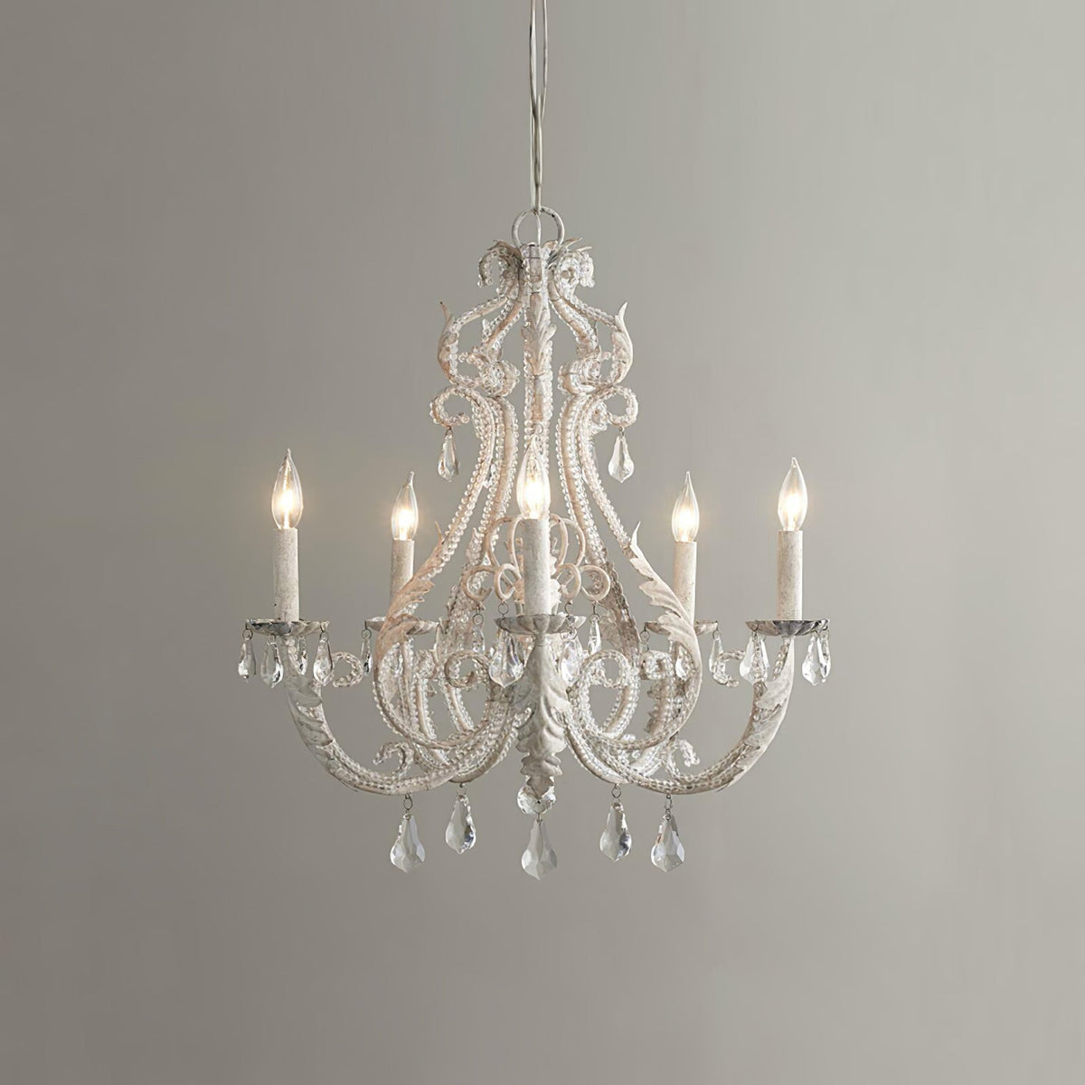 A Clérachic chandelier with unlit candle sleeves, beaded white scrollwork and crystal drops, against a mid-grey wall.