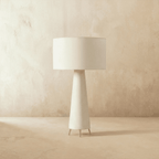 Clérachic unlit white drum shade on a tapering cream base with three slim metal feet, on a pale limewash studio floor.