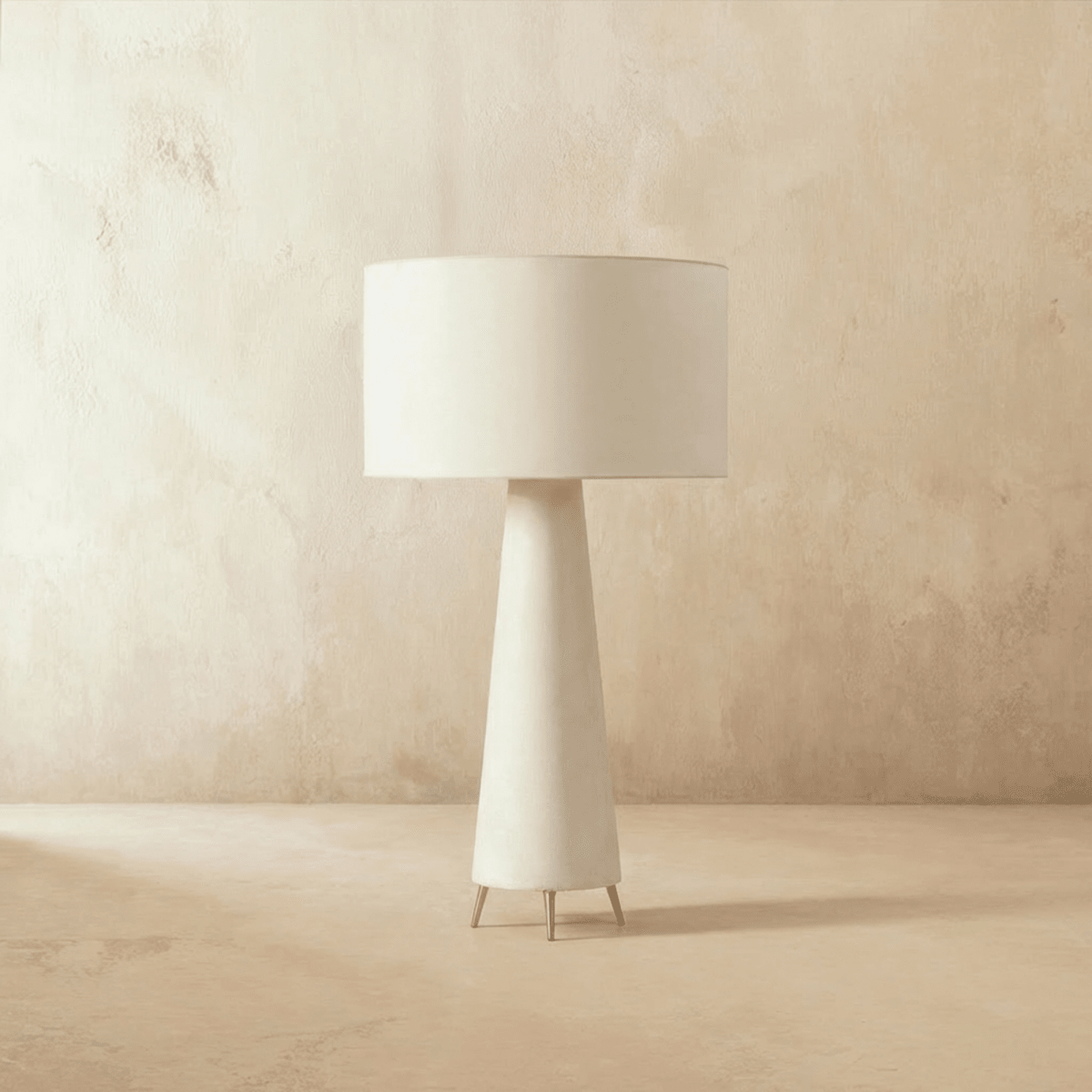 Clérachic unlit white drum shade on a tapering cream base with three slim metal feet, on a pale limewash studio floor.
