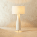 Clérachic larger white linen drum shade lit warm above a tapering cream base and three slim metal feet.