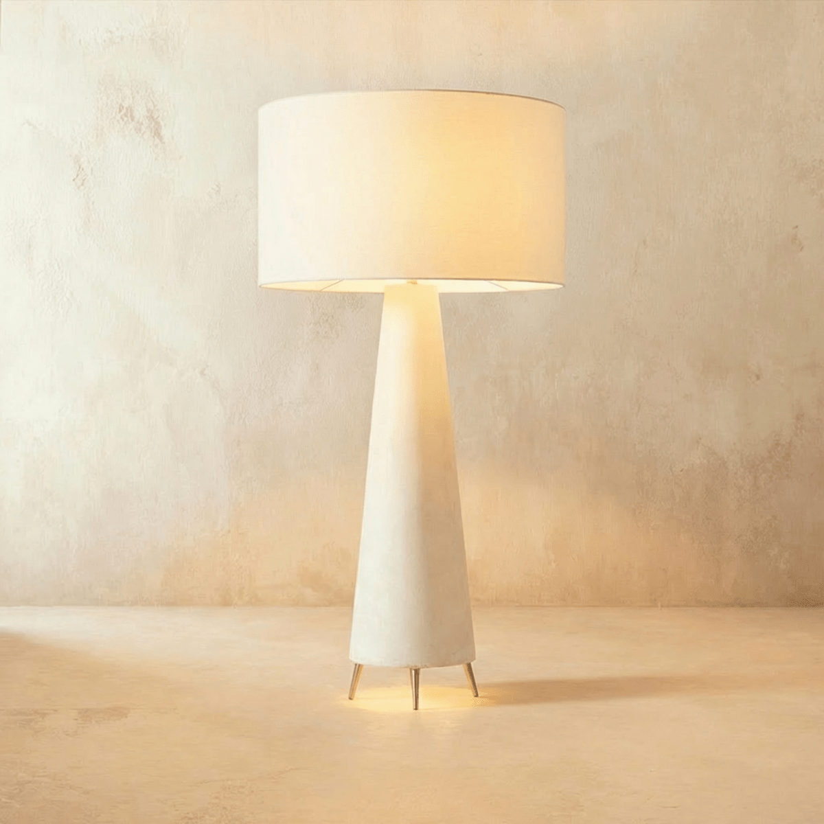 Clérachic tall lamp with a wide white drum shade lit from within, above a tapering cream base on three pointed metal feet.