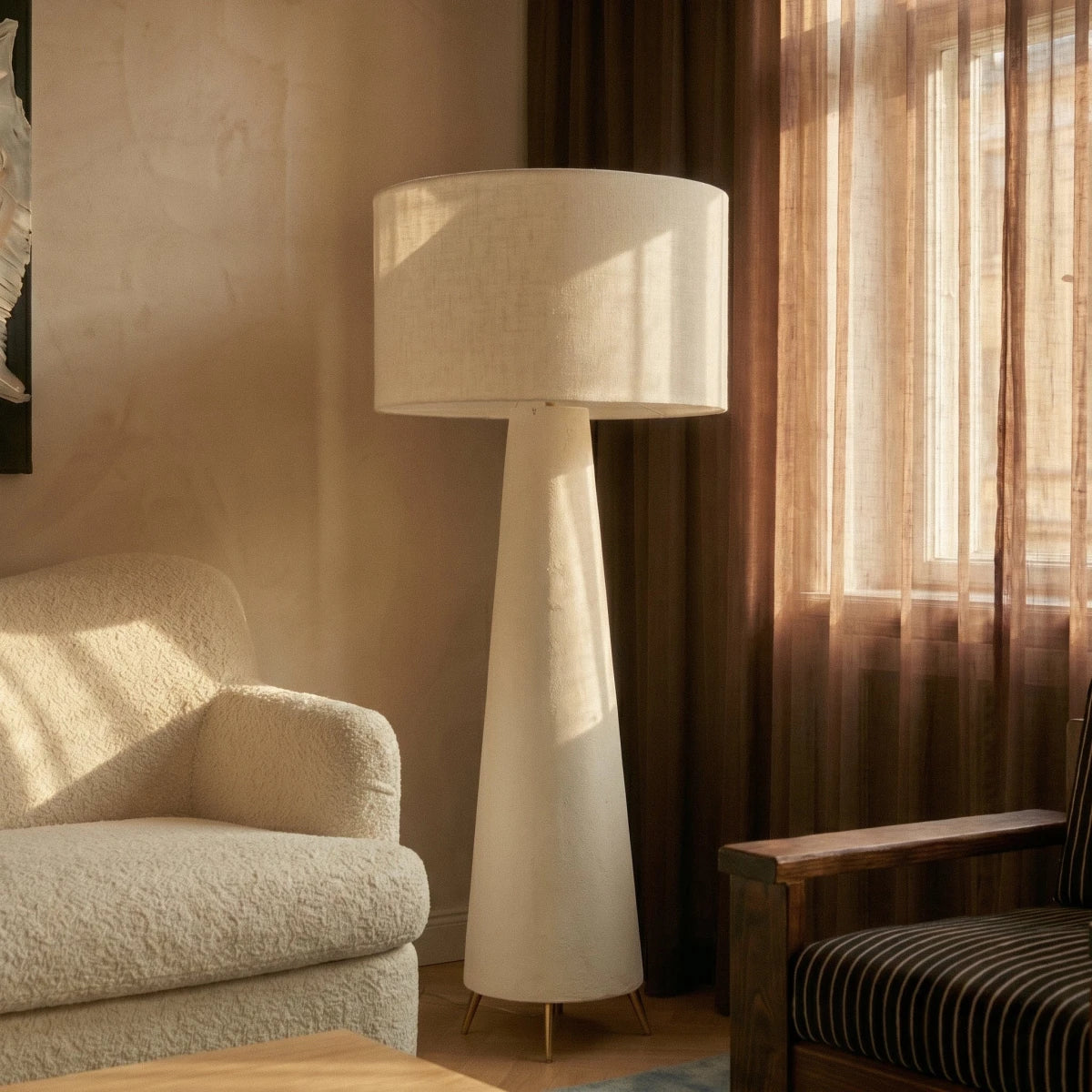 Clérachic tall floor lamp by a sheer-curtained window, white linen drum shade and tapering base beside a bouclé armchair.