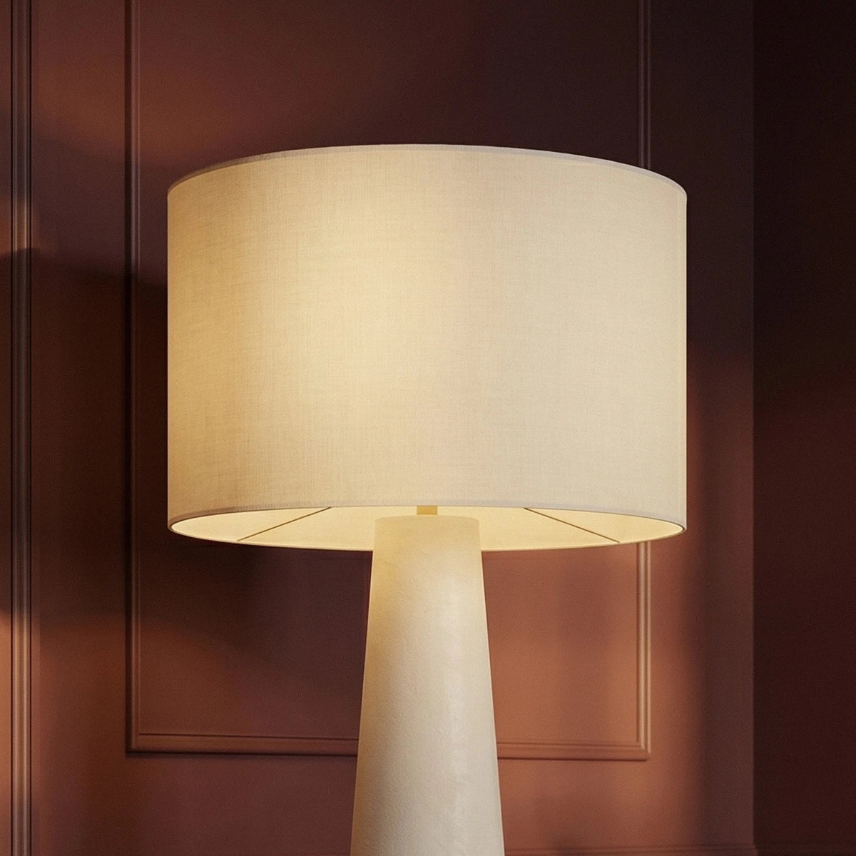 Clérachic close view of a white linen drum shade lit from within, resting on the tapering cream base against plum panelling.