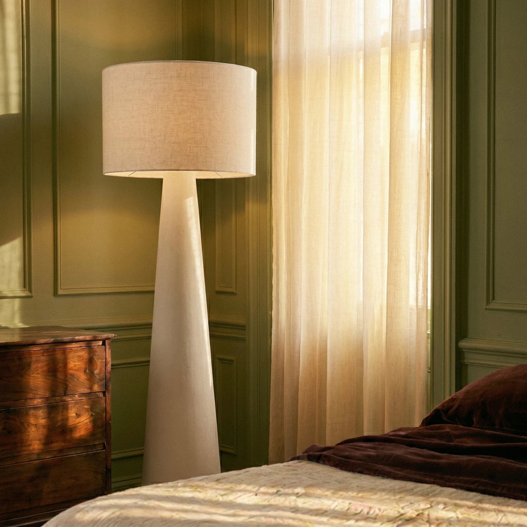 Clérachic tall white linen drum shade floor lamp on a tapering cream base, beside a sheer curtain and a walnut chest of drawers.