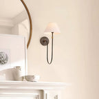 A black Clérachic wall light with a white shade on a cream wall above a white mantel, beside a round mirror and framed prints.