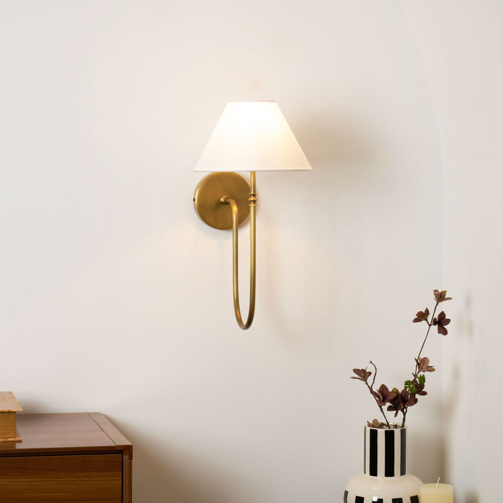 Brass Clérachic wall light with a white fabric shade on a pale wall, above a wooden cabinet and a striped vase of dark stems.